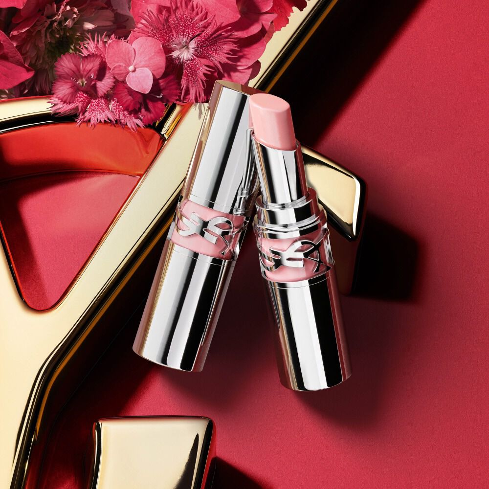 YSL Loveshine lipstick shade 44 in silver packaging, shown open with a soft pink bullet beside the closed tube, styled with pink flowers on a red background with gold YSL logo detail.