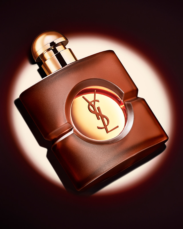 Frosted brown square YSL Opium perfume bottle with its gold cap, displayed at a diagonal angle under a sharp circular spotlight.