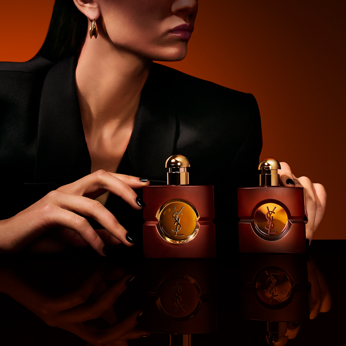 Two square frosted-glass YSL Opium perfume bottles resting on a reflective black surface, framed by a model's hands with black manicured nails against a warm amber background.