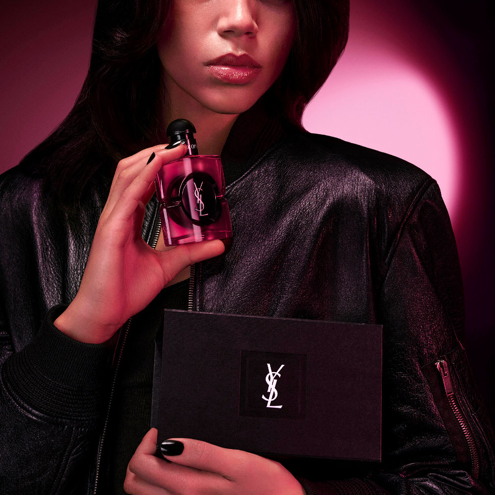 Close-up of a women's model holding Black Opium Eau De Parfum Pink Glaze bottle and a black YSL Beauty gift set in an edgy night setting.