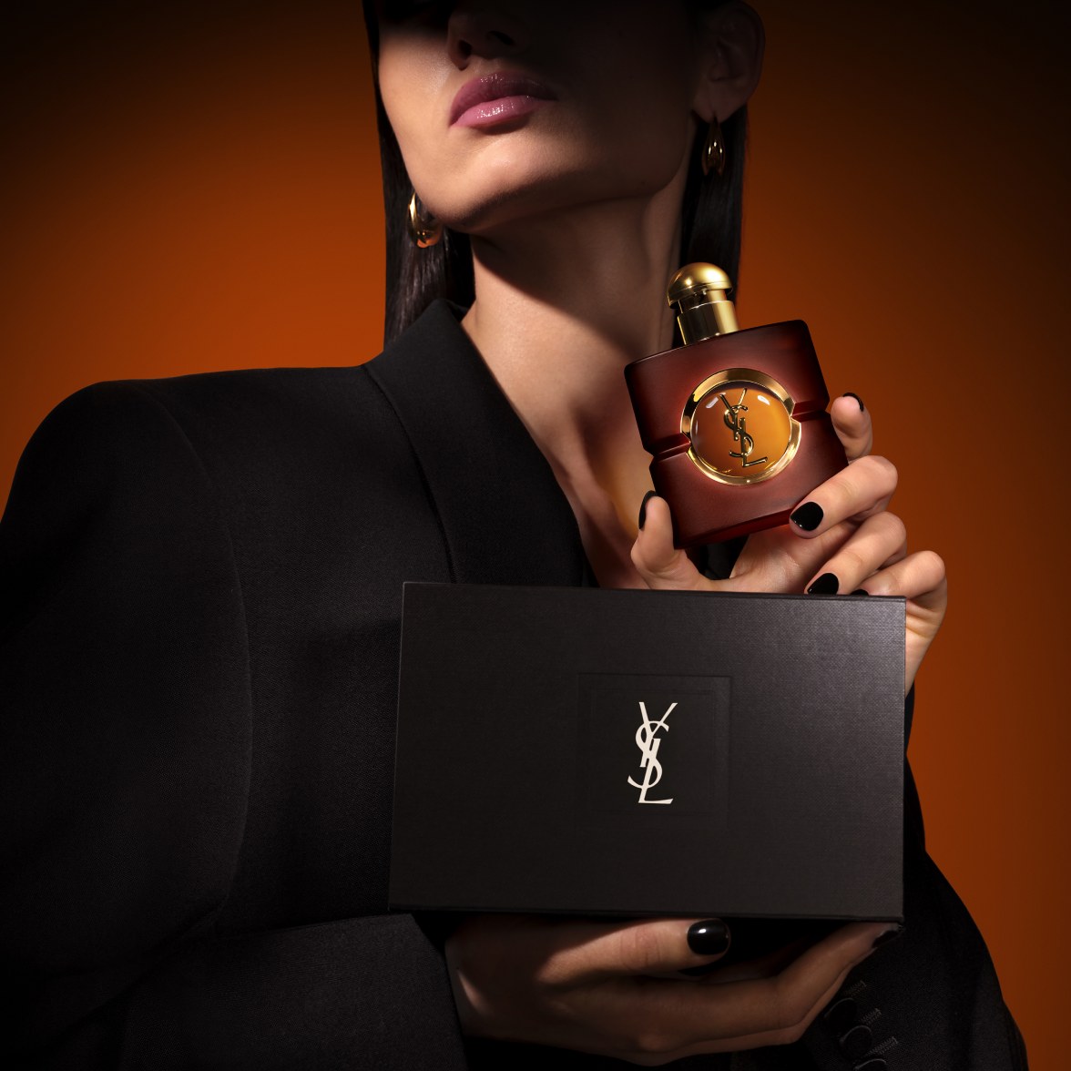 Model in a black blazer holding a square frosted YSL Opium perfume bottle with a round gold cap, resting next to a sleek black textured gift box featuring a silver YSL logo.