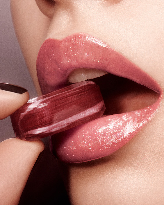 Close-up of glossy pink lips, holding a red candy, showcasing YSL Loveshine's allure.