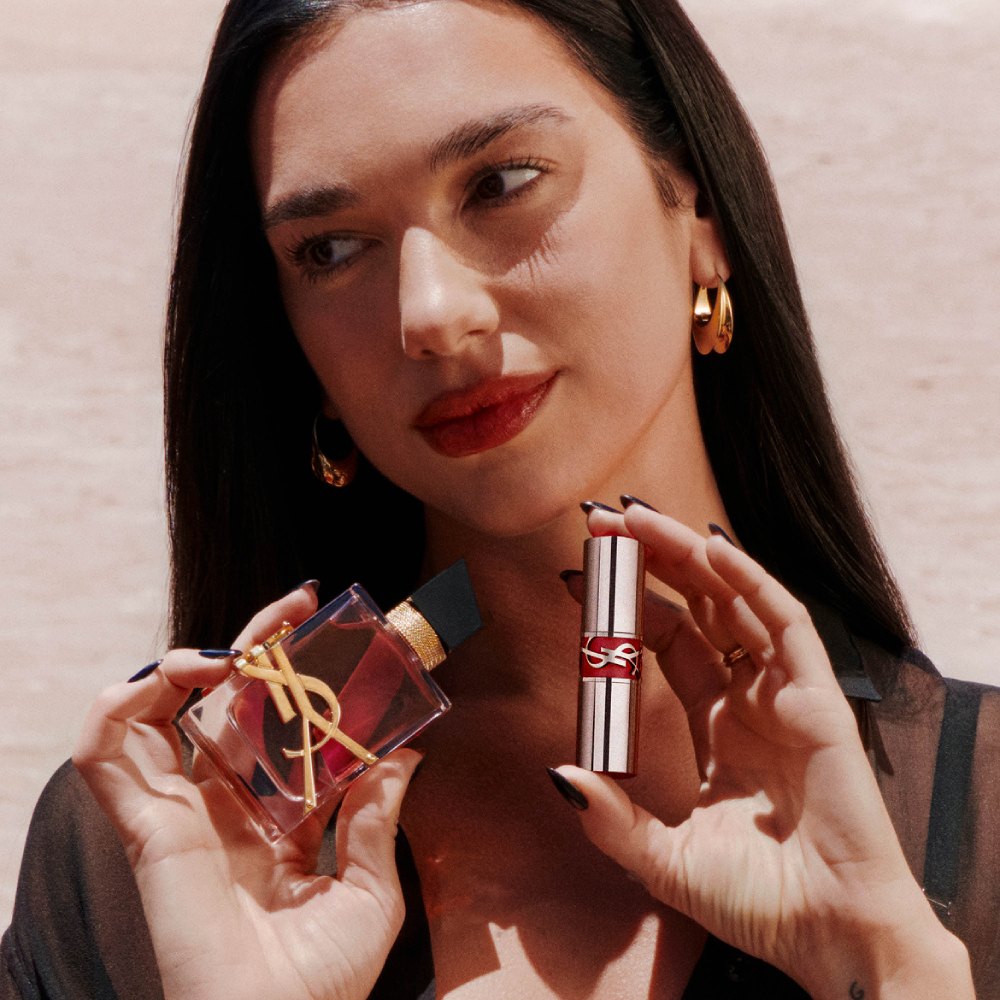 Dua Lipa holdsLibre Eau de parfum Berry Crush and a YSL Loveshine, with red lips and gold earrings.