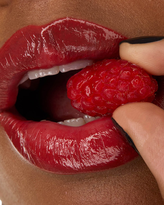Close-up of model Zahara's lips, adorned with YSL LOVESHINE Lipstick in Berry Crush, shade 219, holding a fresh raspberry.