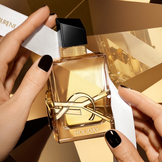 Libre Eau de Parfum perfume bottle with custom engraving, held by hands with black nail polish, on a golden background.