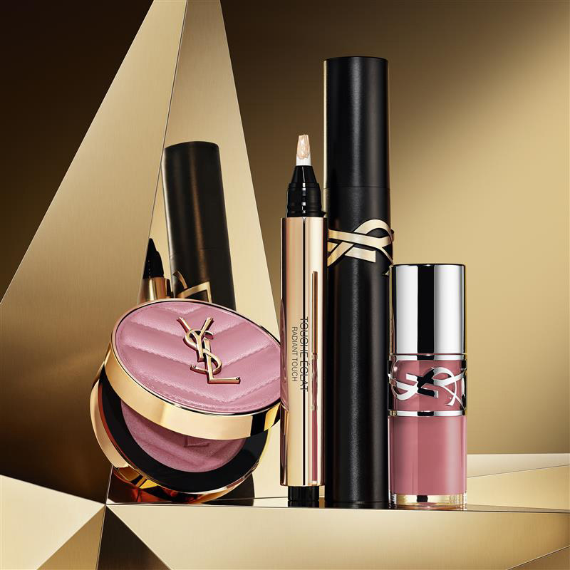 Make Me Blush powder in shade 44, Stylo Touch Eclat, Lash Clash mascara, and YSL Loveshine Plumping Lip Oil Gloss shade 12, on a golden geometric background.