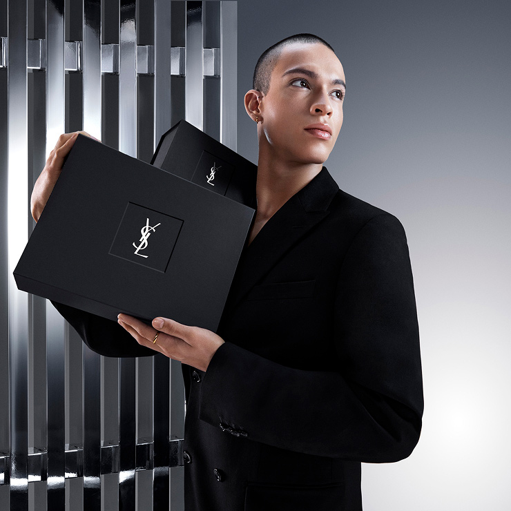 Pierre-Antoine, YSL Beauty model, holding two black YSL gift boxes with silver YSL logos, against a modern grey background.