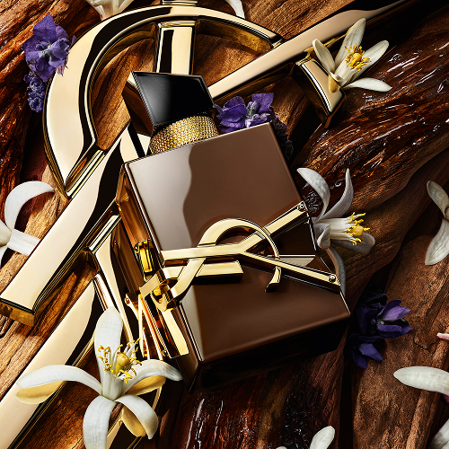 The YSL Cassandre gold rests on raw sandalwood, adorned with white orange blossoms and lavender, with text evoking the sensuality, vibration and energy of sandalwood.