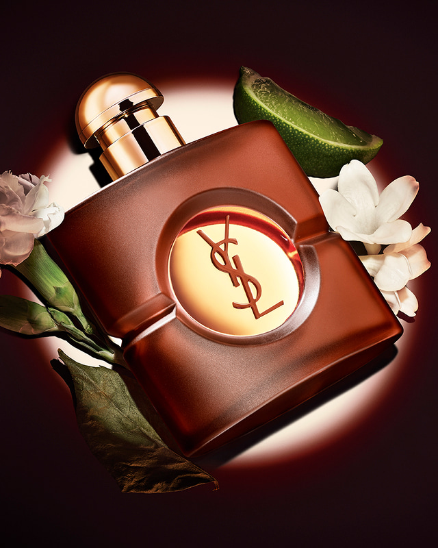 Square frosted brown YSL Opium perfume bottle with a round gold cap, displayed under a spotlight surrounded by a lime wedge, a dried leaf, and white carnation flowers.