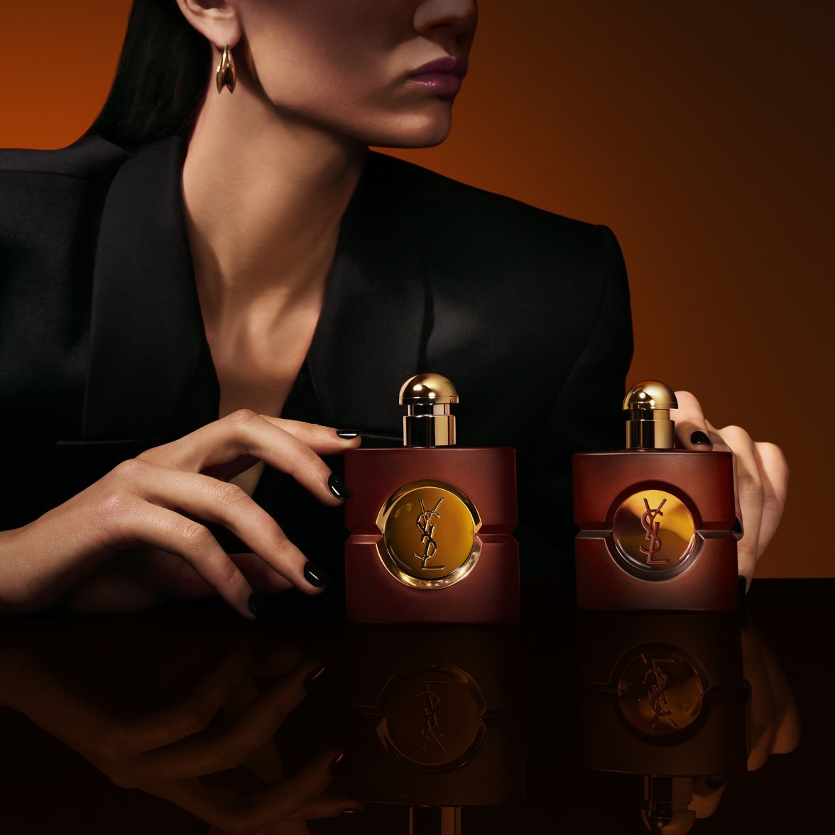 Two square frosted-glass YSL Opium perfume bottles resting on a reflective black surface, framed by a model's hands with black manicured nails against a warm amber background.
