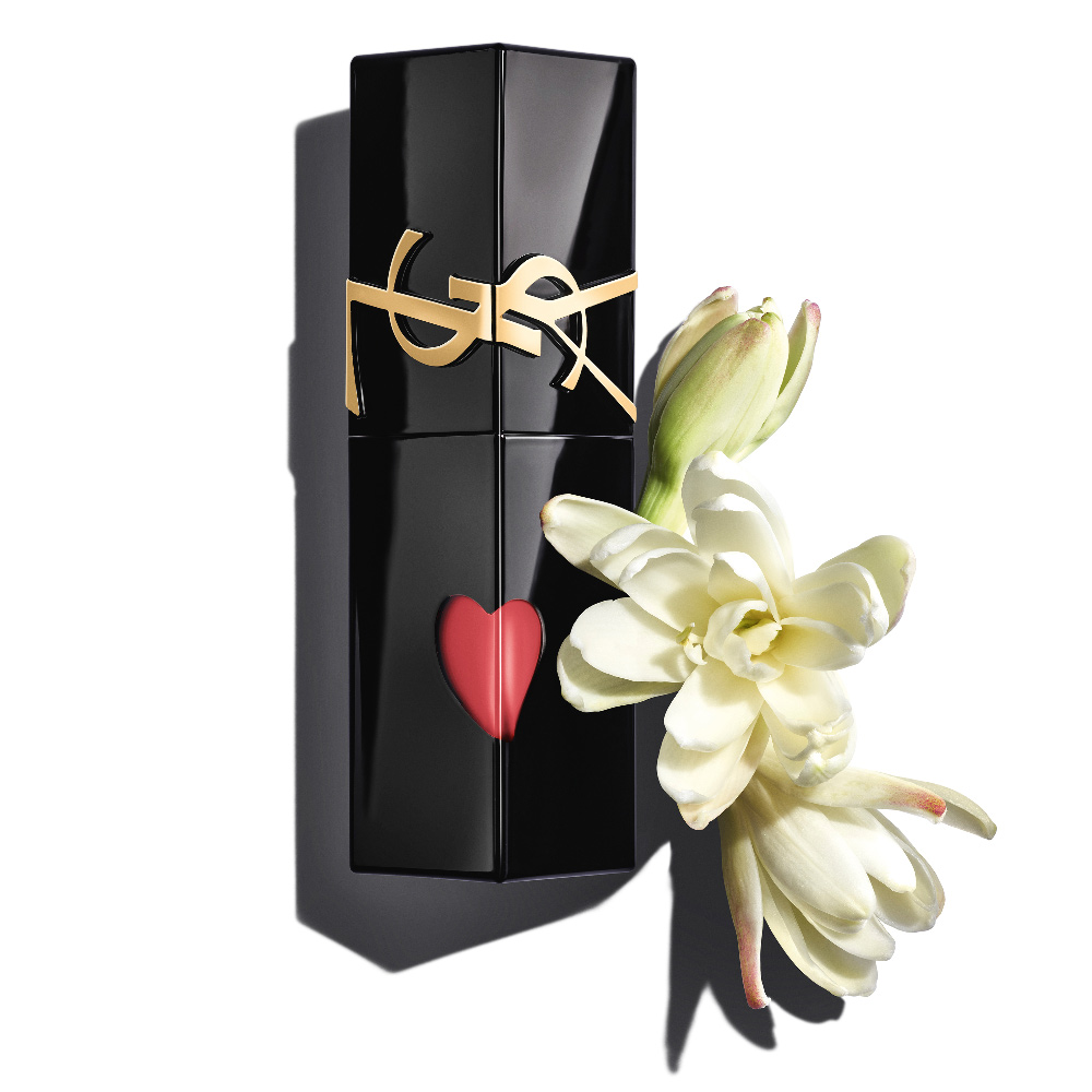 A glossy black YSL lip stain bottle with a red heart-shaped cutout, elegantly displayed next to delicate, blooming white tuberoses.
