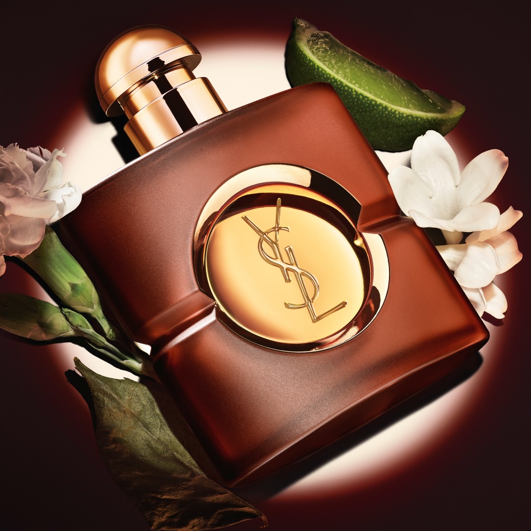 Square frosted brown YSL Opium perfume bottle with a round gold cap, displayed under a spotlight surrounded by a lime wedge, a dried leaf, and white carnation flowers.