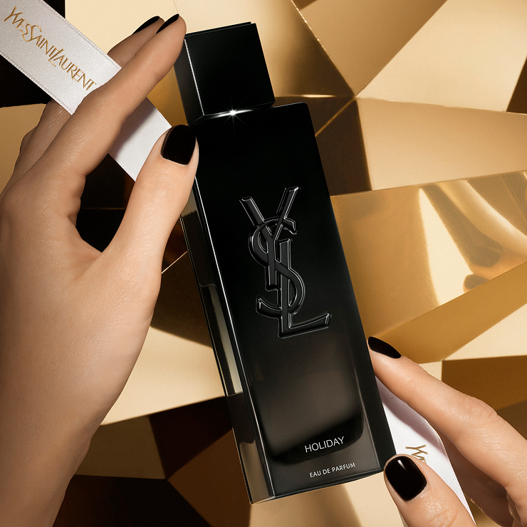 Hands holding a MYSLF Eau de Parfum bottle with "HOLIDAY" engraved, against a golden geometric background.