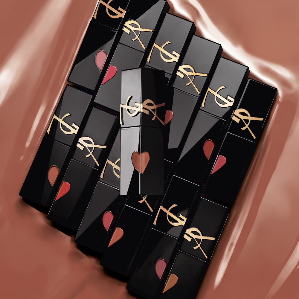 Multiple glossy black YSL lip stain bottles featuring heart-shaped cutouts and gold logos, neatly arranged in rows on a shiny, caramel-nude liquid backdrop.