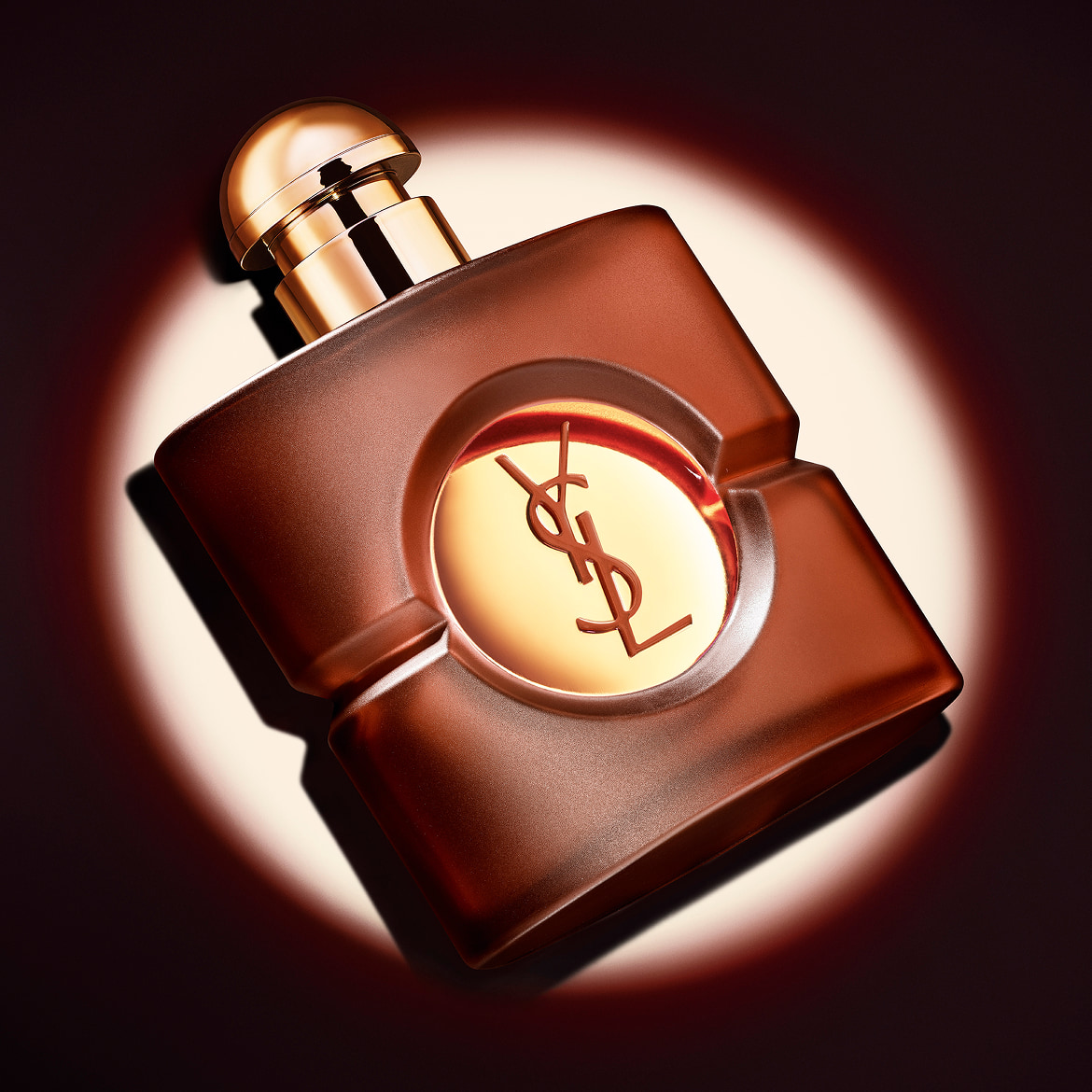Frosted brown square YSL Opium perfume bottle with its gold cap, displayed at a diagonal angle under a sharp circular spotlight.