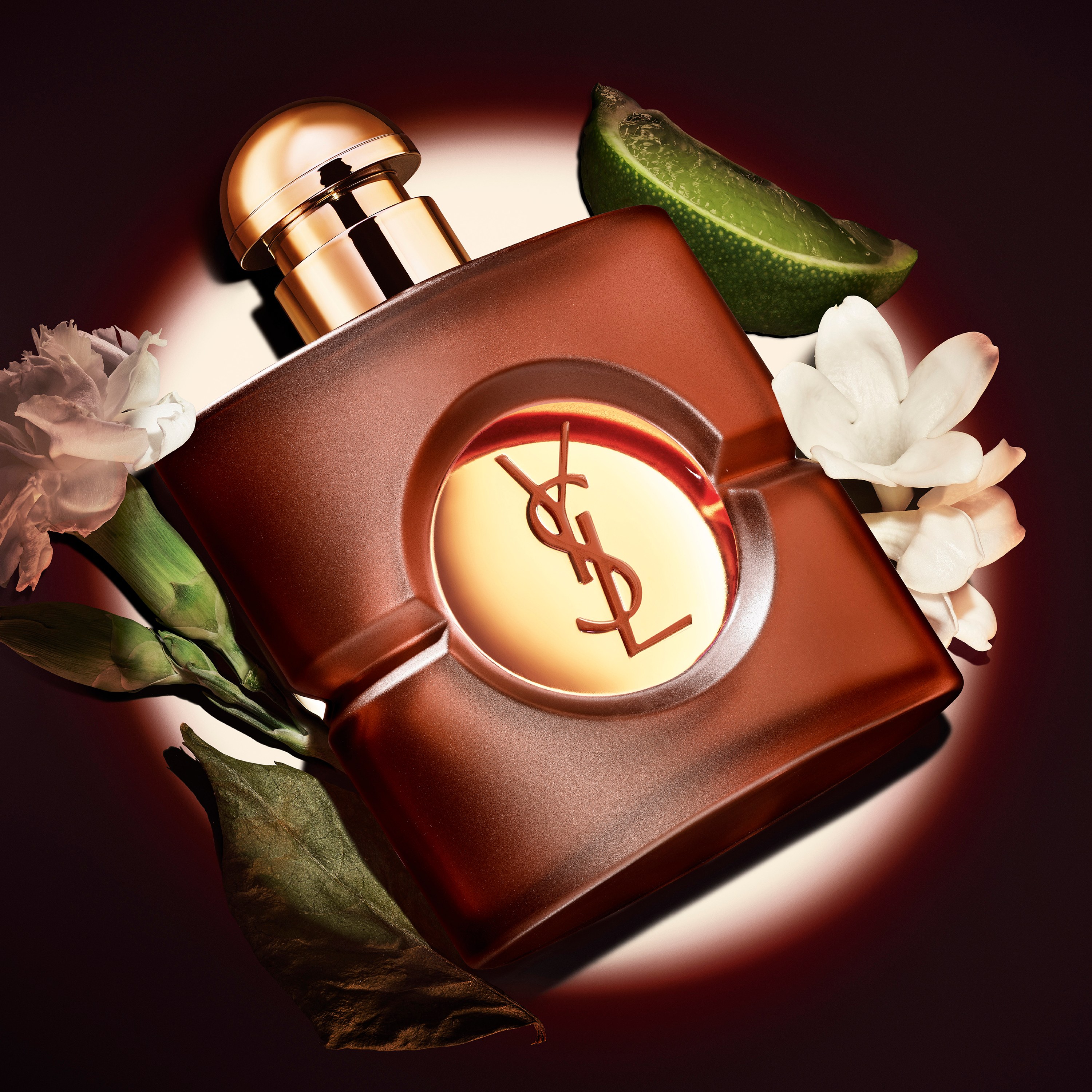 Square frosted brown YSL Opium perfume bottle with a round gold cap, displayed under a spotlight surrounded by a lime wedge, a dried leaf, and white carnation flowers.