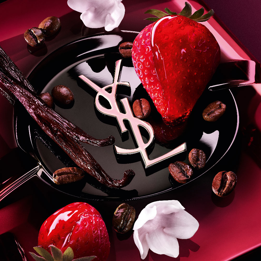 Close-up of fresh strawberries, vanilla pods, jasmine flowers, and roasted coffee beans arranged over the Black Opium Eau de Parfum Pink Glaze bottle with the iconic YSL cassandre.