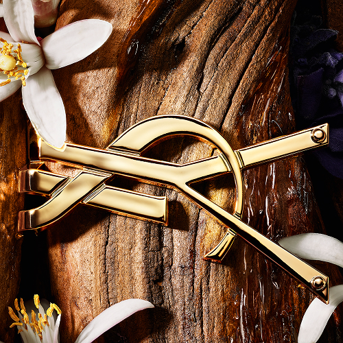 Golden YSL Cassandre lying on raw sandalwood bark with white orange blossoms.