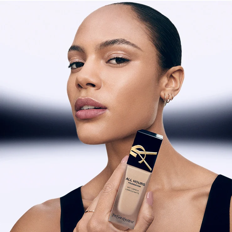 Model with medium skin tone holding YSL All Hours Foundation bottle, wearing a black top, close-up view.