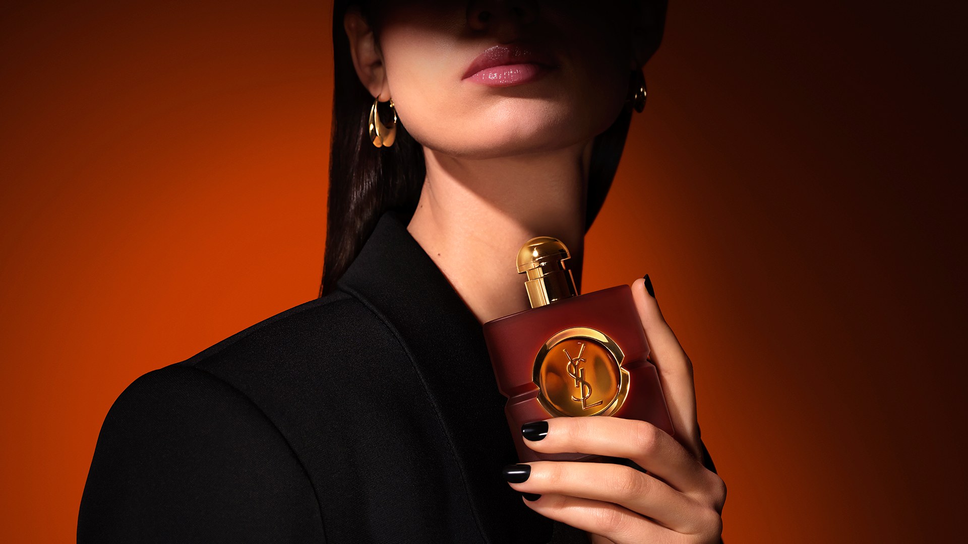 Model with a glossy pink lip wearing a black blazer and gold earrings, holding a square frosted brown YSL Opium perfume bottle against her neck, set against a warm amber-orange background.