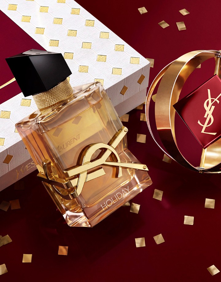YSL Libre perfume bottle with HOLIDAY engraved at the bottom, featuring a bold gold logo resting against a white and gold diamond-patterned gift box with gold confetti on a deep red surface.